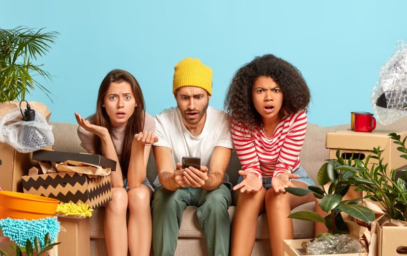 Three mixed race friends pose together on comfortable couch, have frustrated puzzled expressions, surf internet on mobile phone, cannot find appropriate interior for new house move in bought apartment
