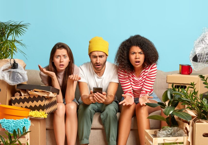 Three mixed race friends pose together on comfortable couch, have frustrated puzzled expressions, surf internet on mobile phone, cannot find appropriate interior for new house move in bought apartment